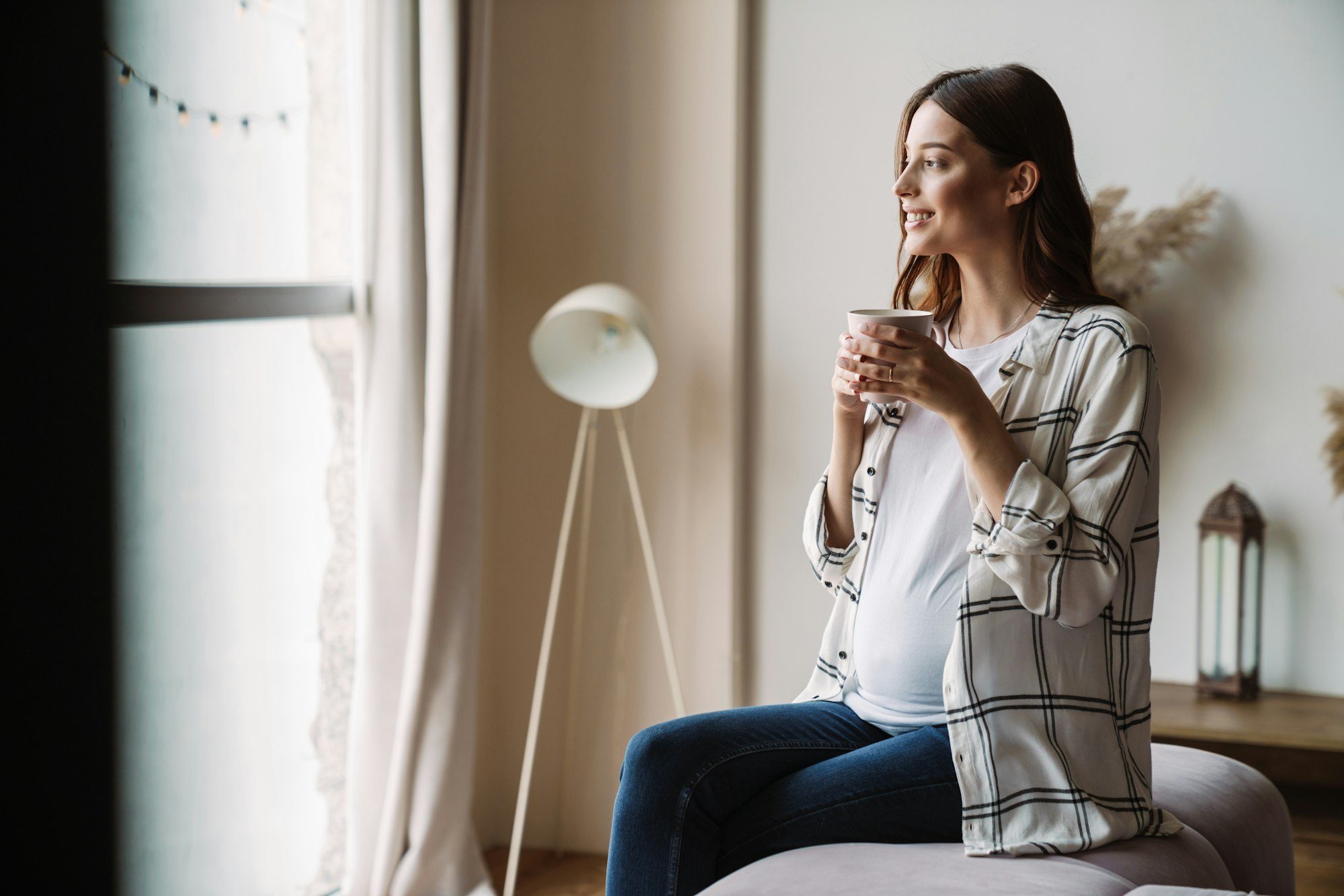 Beautiful happy pregnant woman smiling and drinking coffee while sitting on couch