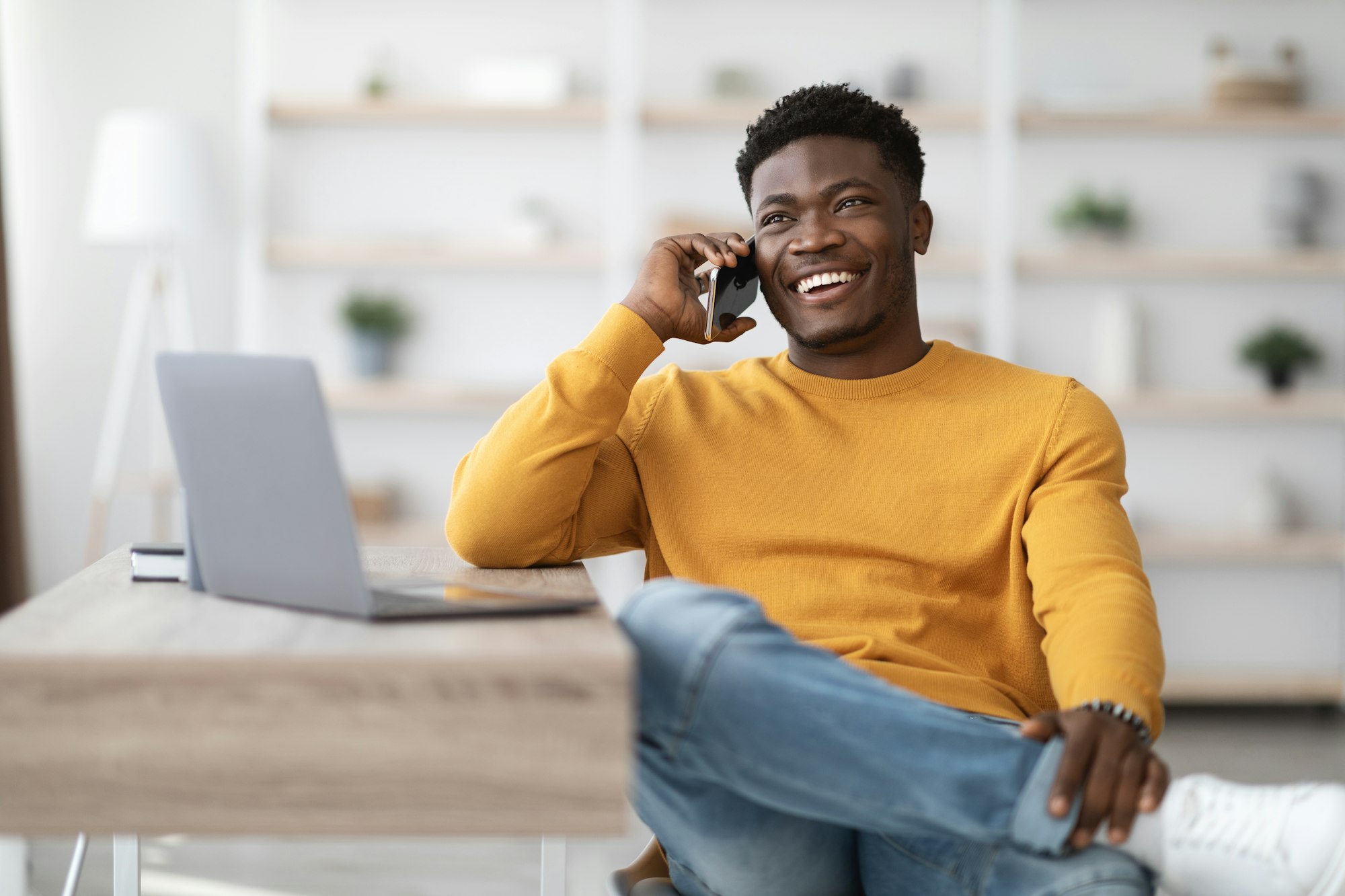 Cheerful man sitting at table with laptop, talking on phone