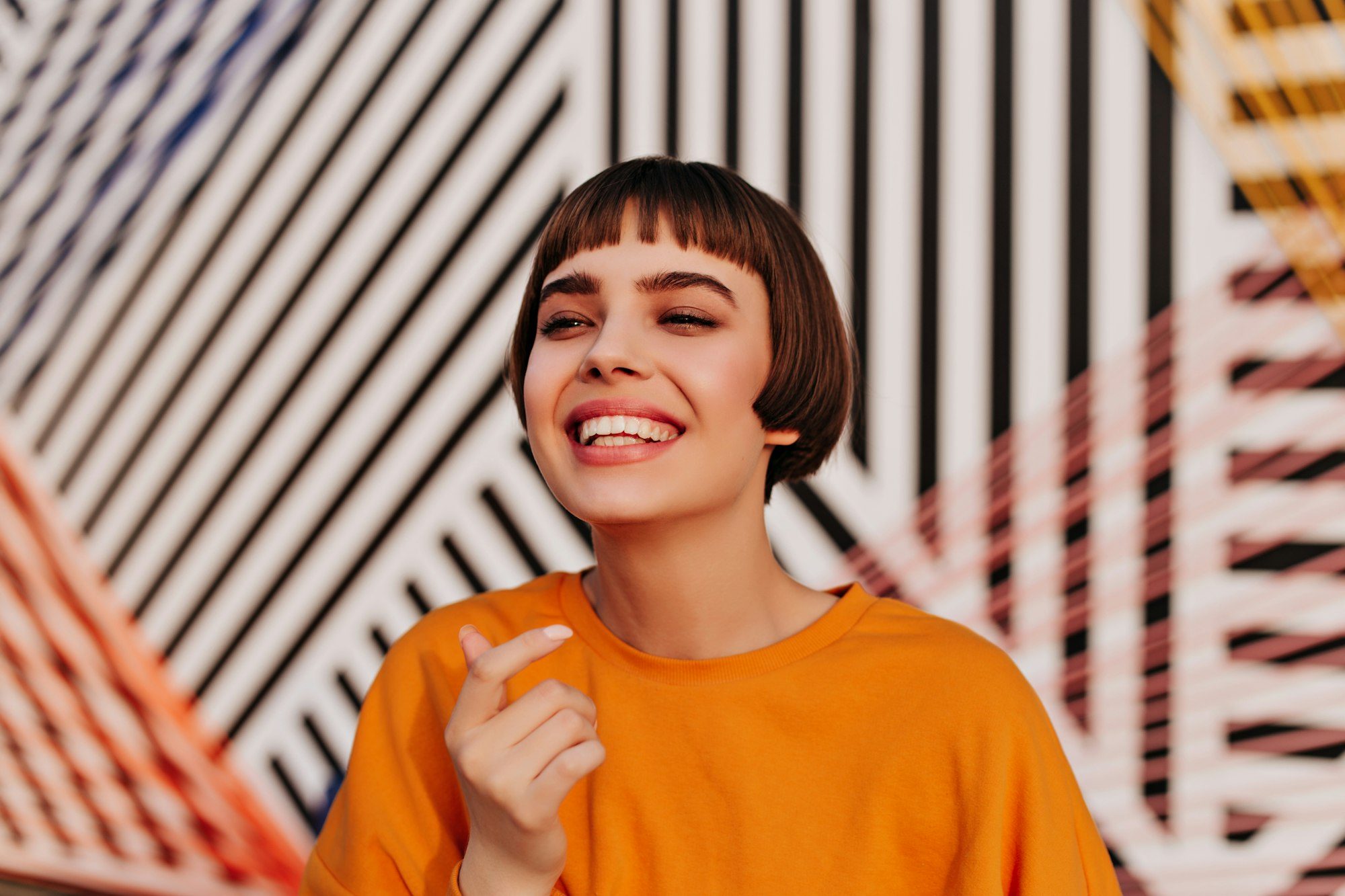 Optimistic teen girl in orange sweatshirt smiling on striped backdrop. Brunette woman in bright out